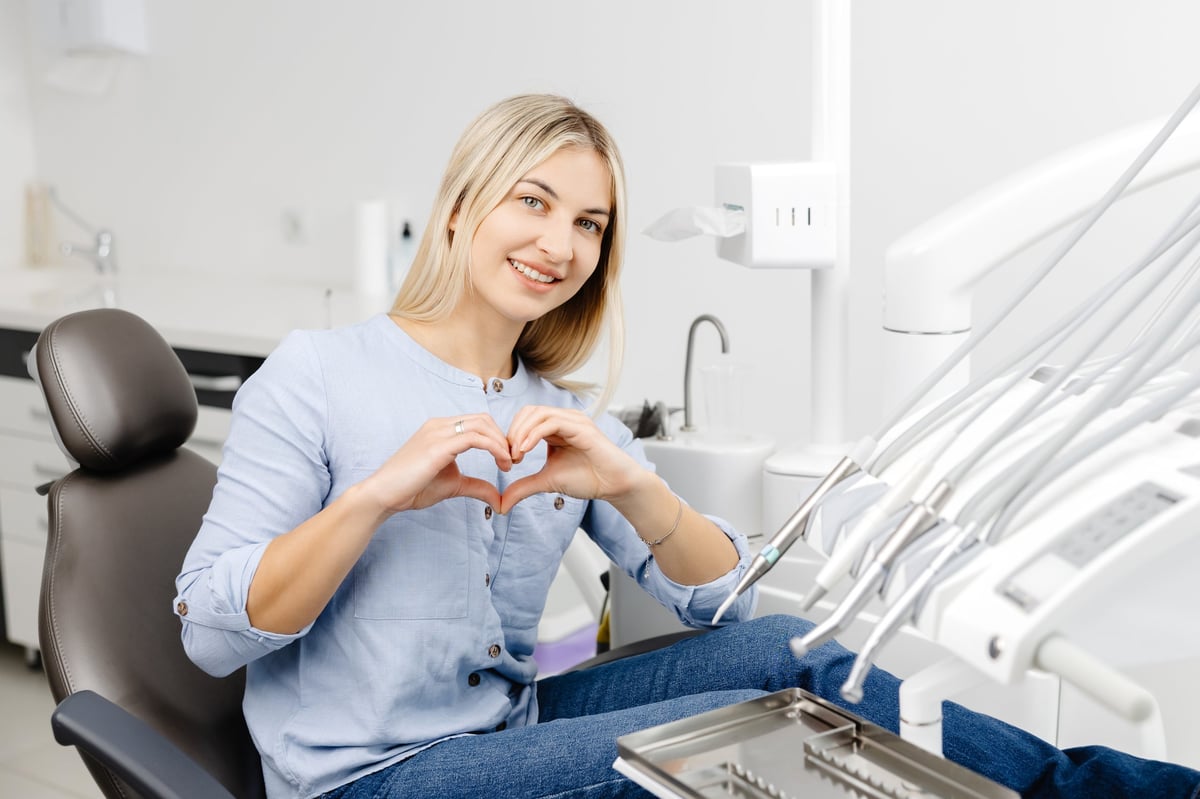 Young woman making heart with her hands sitting in a dental chair, showing love and appreciation for dental care