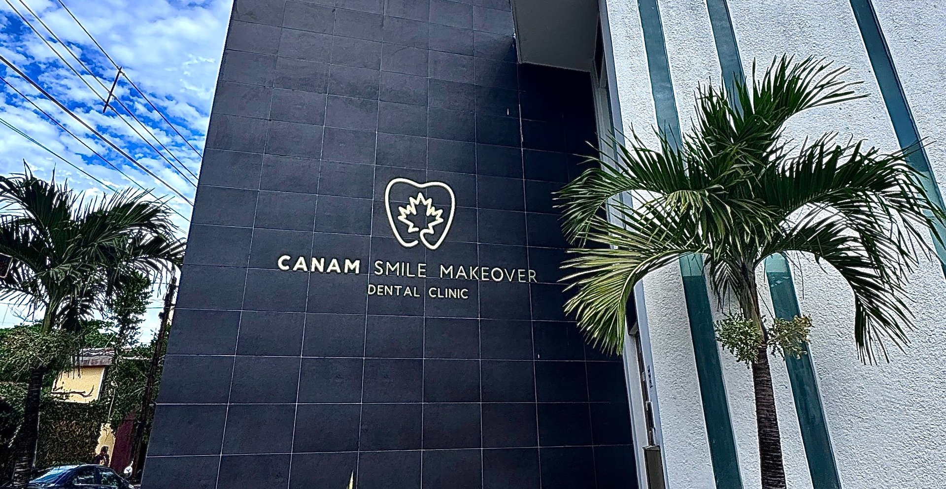 Modern dental clinic building with Canam Smile Recover logo on dark facade, surrounded by palm trees and clear blue sky