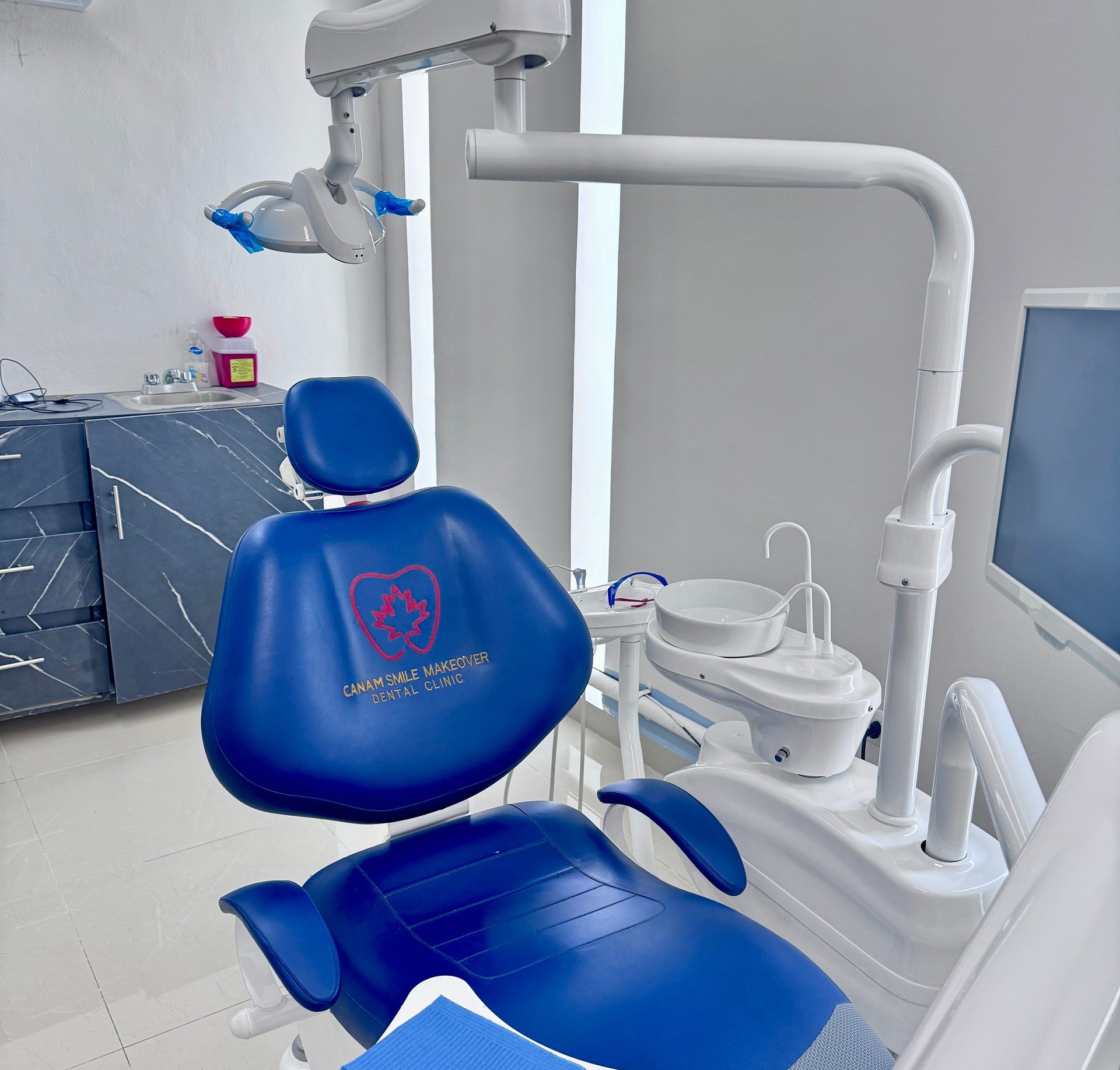 Modern dental clinic treatment room with blue patient chair, overhead lamp, white suction equipment, and monitor in background