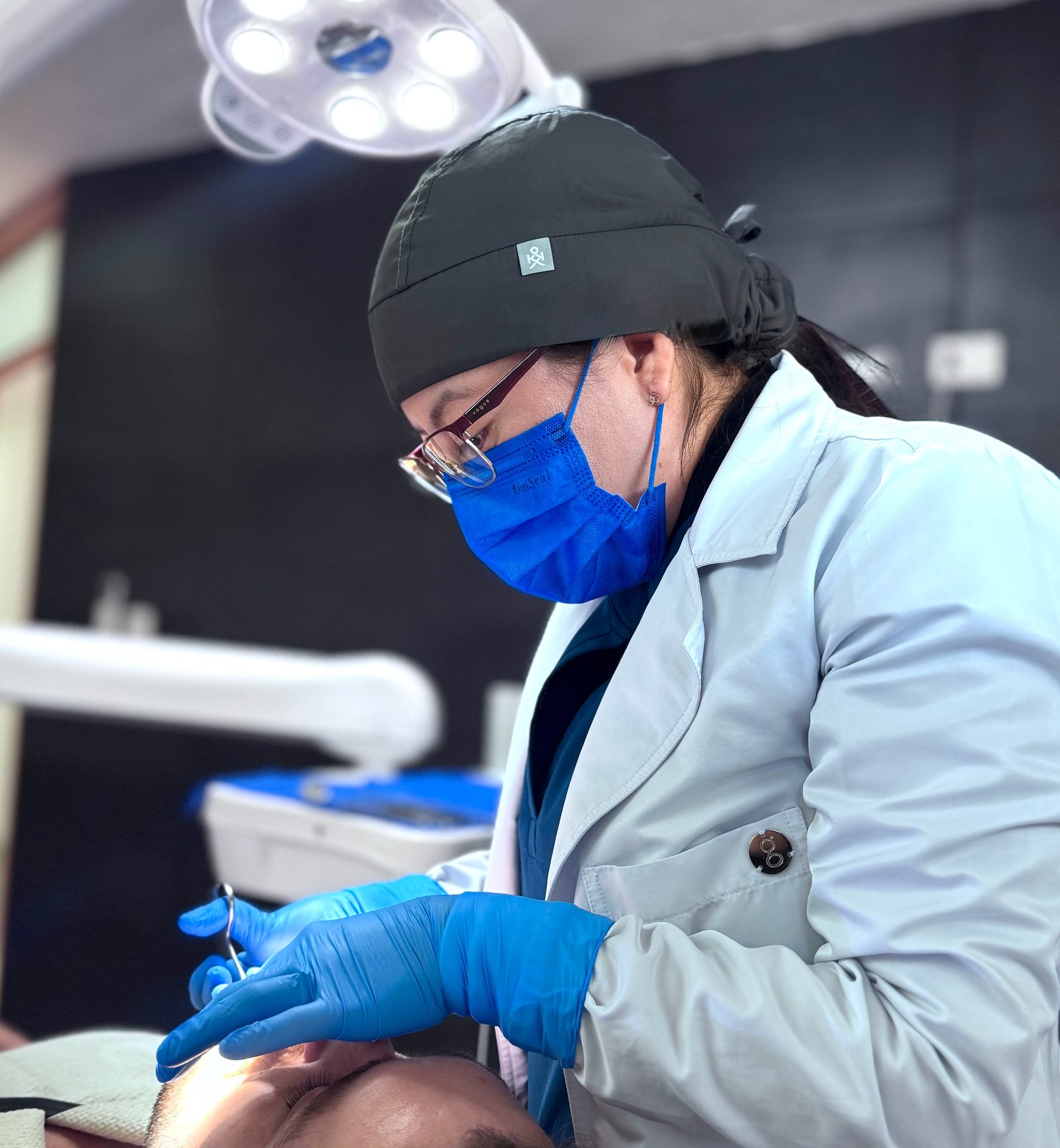 Dental professional in white coat, blue mask and gloves examining patient under overhead surgical light in clinical setting