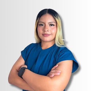 Retrato de mujer con cabello rubio y oscuro usando playera azul, brazos cruzados, contra fondo blanco.