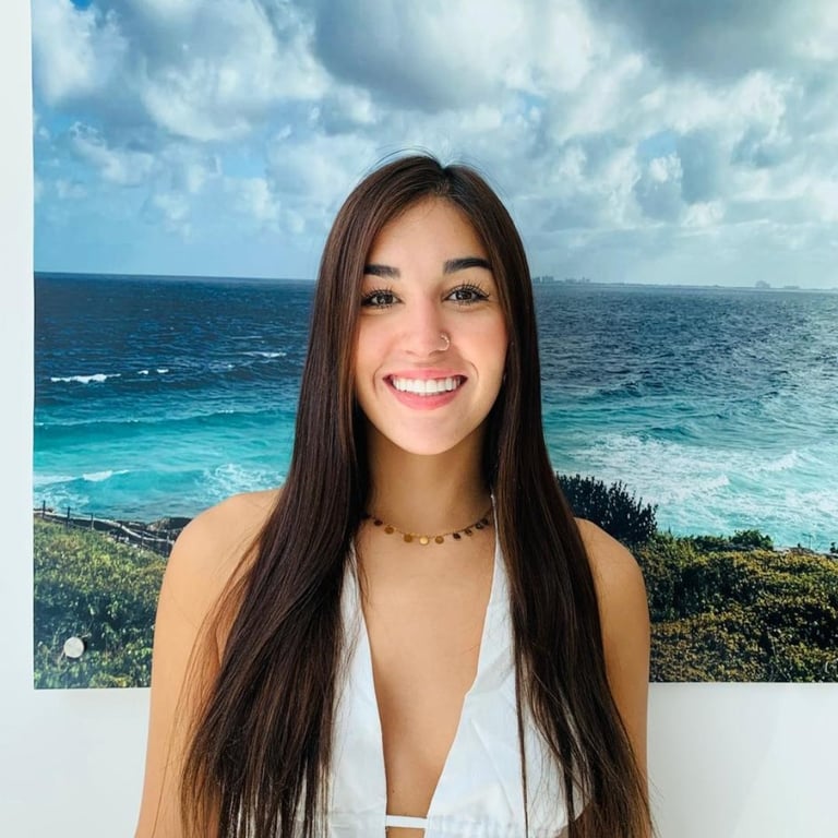 Smiling woman with long dark hair wearing a white crop top and beige cardigan against a beach backdrop with turquoise water